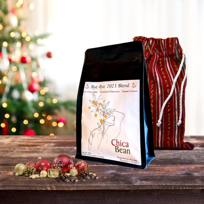 A bag of coffee called Bye-Bye 2023 Blend in front of another bag of coffee that's inside a colorful fabric bag with a Christmas tree blurred in the background