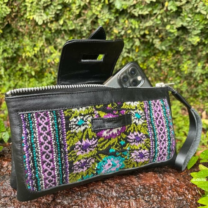 A phone sticking out of a black leather clutch with purple, blue, and green traditional Guatemalan Fabric sown into it. Closes with a latch and has a wristband