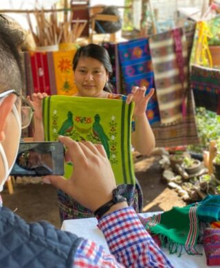 A woman holding up a green pillow case that she wove. The pillow case has 2 birds (the Guatemalan Quetzal) and there is a man taking a picture of her with a cellphone.