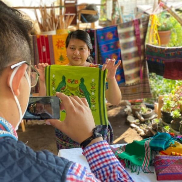 A woman holding up a green pillow case that she wove. The pillow case has 2 birds (the Guatemalan Quetzal) and there is a man taking a picture of her with a cellphone.